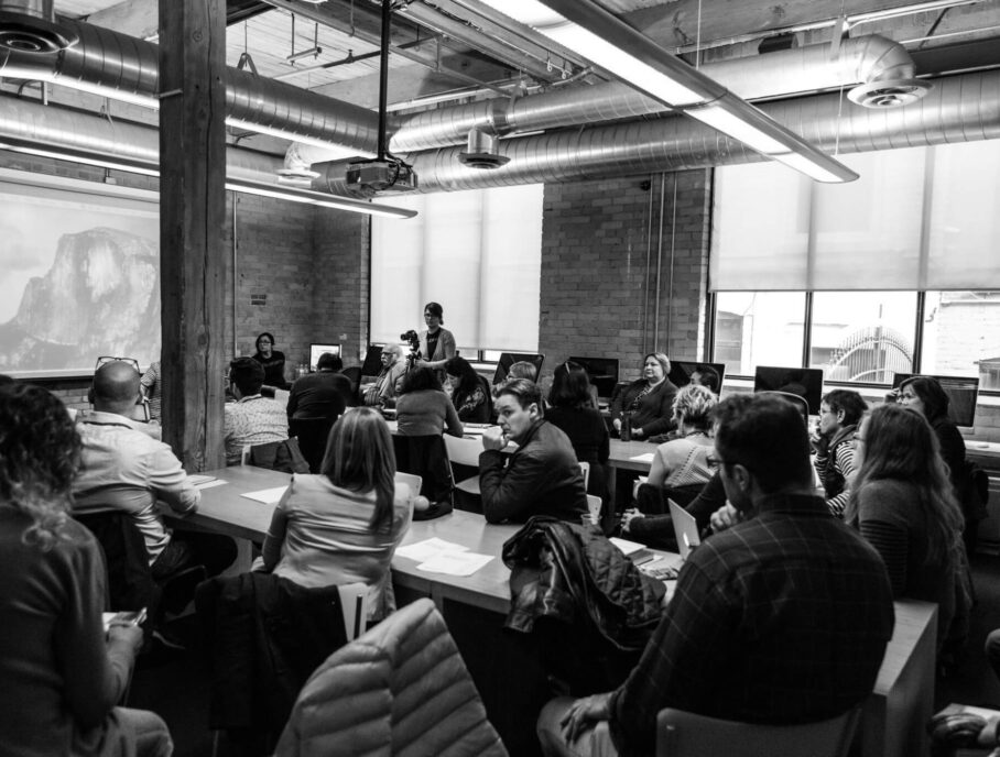 A black and white image of a large group of people sitting in a conference room, watching a presentation.